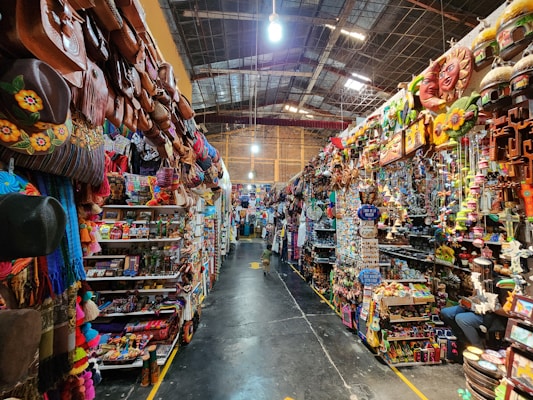 A vibrant, bustling indoor market stall filled with an array of colorful merchandise. The shelves are stocked with leather bags, hats, crafts, and various decorative items. The scene is lively, with bright colors and diverse items creating a visually rich environment.