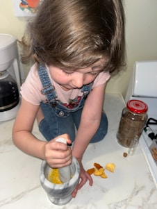 A child wearing a pink shirt and denim overalls is using a marble mortar and pestle. They are grinding yellow spices inside the mortar on a white countertop. Nearby, there are spice jars and an assortment of ingredients.