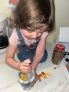 A child wearing a pink shirt and denim overalls is using a marble mortar and pestle. They are grinding yellow spices inside the mortar on a white countertop. Nearby, there are spice jars and an assortment of ingredients.