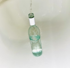 A clear glass bottle standing upright in a white sink as water flows into it from above. The bottle is partially filled with water, and reflections from the glass create gentle ripples on the surface.