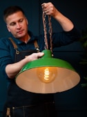A person wearing a dark apron is holding a hanging light fixture with a green shade. The fixture has a decorative, glowing filament bulb inside. The person appears focused, adjusting or holding the chain of the lamp.
