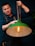 A person wearing a dark apron is holding a hanging light fixture with a green shade. The fixture has a decorative, glowing filament bulb inside. The person appears focused, adjusting or holding the chain of the lamp.