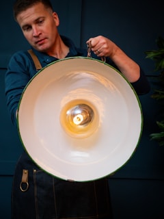 A person holding a large, industrial-style pendant light fixture. The light is turned on, emitting a warm glow. They are wearing a dark apron over a blue shirt. The background appears dark, contrasting with the bright light.