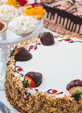 A variety of desserts including a layered cake covered in sliced almonds and decorated with chocolate-covered strawberries. Nearby are fruit tarts topped with fresh fruits like kiwi and strawberries as well as a meringue-topped pie.