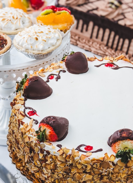 A variety of desserts including a layered cake covered in sliced almonds and decorated with chocolate-covered strawberries. Nearby are fruit tarts topped with fresh fruits like kiwi and strawberries as well as a meringue-topped pie.