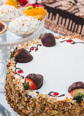 A variety of desserts including a layered cake covered in sliced almonds and decorated with chocolate-covered strawberries. Nearby are fruit tarts topped with fresh fruits like kiwi and strawberries as well as a meringue-topped pie.