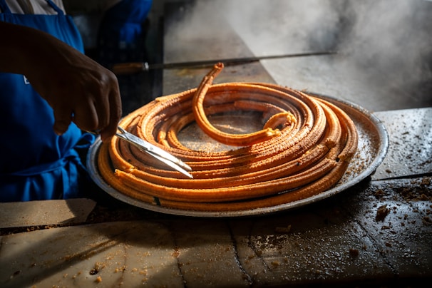 A person wearing a blue apron is slicing a large circular coil of churros on a metal tray. The tray is set on a surface with visible crumbs, and there is steam in the background, suggesting the churros are hot and freshly made.