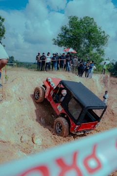 A red off-road vehicle is climbing a steep, muddy terrain while a group of spectators watches from the sidelines. One spectator is holding a colorful umbrella. The background features a tree and a cloudy sky.