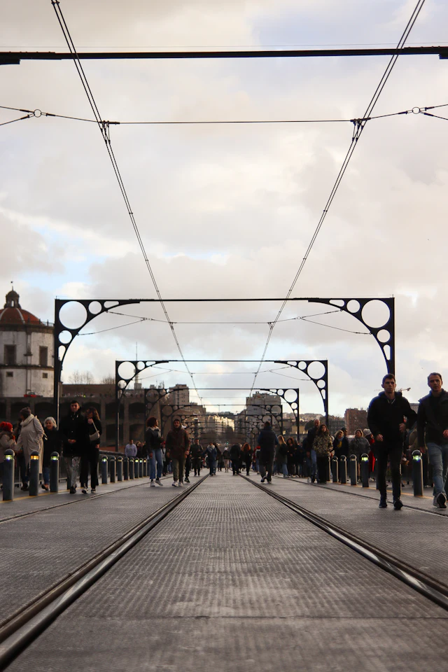 Pedestrians crossing the upper deck of the Dom Luís I Bridge beneath its iron lattice overhead frames and tram wires.