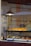 Inside a bakery, various pastries are displayed on a shelf behind a glass window. The shelf is well-lit, highlighting neatly arranged tarts and a selection of sandwiches and croissants. A hanging metal lamp provides additional warm lighting.