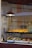 Inside a bakery, various pastries are displayed on a shelf behind a glass window. The shelf is well-lit, highlighting neatly arranged tarts and a selection of sandwiches and croissants. A hanging metal lamp provides additional warm lighting.
