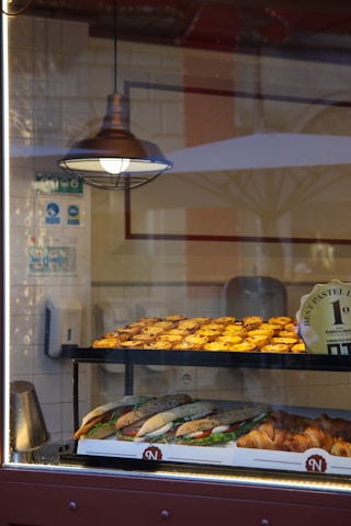 Inside a bakery, various pastries are displayed on a shelf behind a glass window. The shelf is well-lit, highlighting neatly arranged tarts and a selection of sandwiches and croissants. A hanging metal lamp provides additional warm lighting.