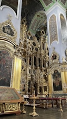 A richly decorated interior space featuring ornate gold embellishments, large paintings, and a grand chandelier. The architecture includes intricate woodwork and detailed carvings. Religious motifs and iconography are prominent.