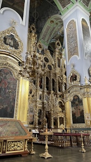 A richly decorated interior space featuring ornate gold embellishments, large paintings, and a grand chandelier. The architecture includes intricate woodwork and detailed carvings. Religious motifs and iconography are prominent.