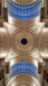 An ornate, detailed ceiling featuring intricate carvings and decorative patterns. There is a large, circular skylight in the center surrounded by elaborate plasterwork. The ceiling is decorated with floral and geometric motifs, and is highlighted by soft lighting. Two sections of gridded glass windows allow natural light to filter through.