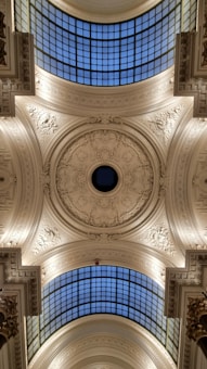 An ornate, detailed ceiling featuring intricate carvings and decorative patterns. There is a large, circular skylight in the center surrounded by elaborate plasterwork. The ceiling is decorated with floral and geometric motifs, and is highlighted by soft lighting. Two sections of gridded glass windows allow natural light to filter through.