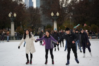 A group of people are ice skating in an outdoor rink surrounded by trees and city buildings. Three individuals in the foreground are holding hands, wearing winter clothing. The background features several other skaters and decorative street lamps.