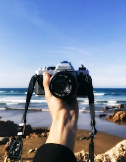 A hand holds a vintage Nikon camera against the backdrop of a sandy beach with waves and rocks. The sky is clear and blue, suggesting a bright and sunny day.