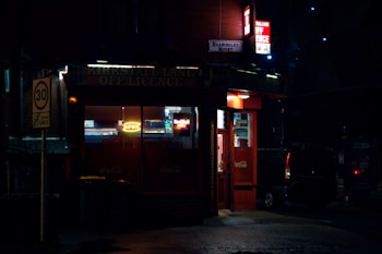 A small off-licence store at night with bright neon lights that illuminate the entrance. The sign above the shop reads 'Kirkstall Lane Off Licence' and another neon sign says 'Open'. The streetlights and shop lights cast a warm glow on the wet pavement. A van is parked nearby, and a 30 mph speed limit sign is visible.