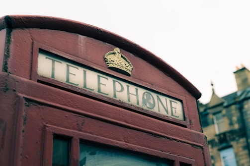 A vintage red telephone box with a crown emblem above the sign, partially in view. The glass is slightly weathered, and the background shows blurred rooftops with a cloudy sky.