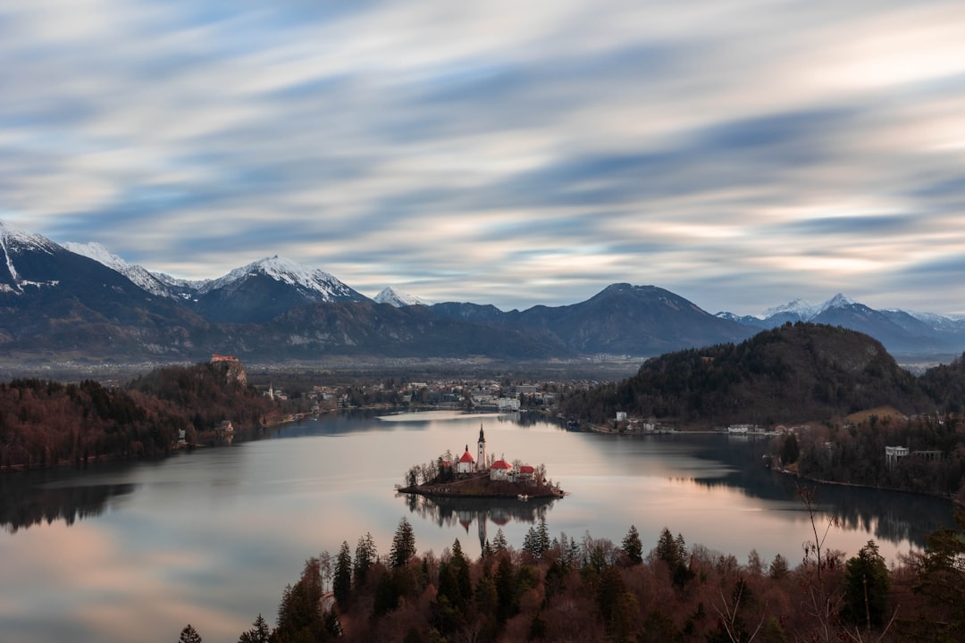 a lake surrounded by trees and mountains under a cloudy sky, Bled lake with the famous island with the church in Sloveni