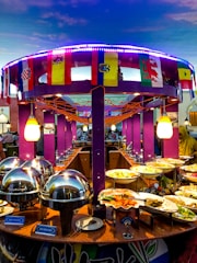 A vibrant buffet setup featuring an array of dishes including seafood and vegetables. The buffet is decorated with international flags, and the setting includes bright purple columns and string lights. Chafing dishes and labeled food containers are prominently displayed.