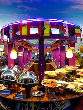 A vibrant buffet setup featuring an array of dishes including seafood and vegetables. The buffet is decorated with international flags, and the setting includes bright purple columns and string lights. Chafing dishes and labeled food containers are prominently displayed.