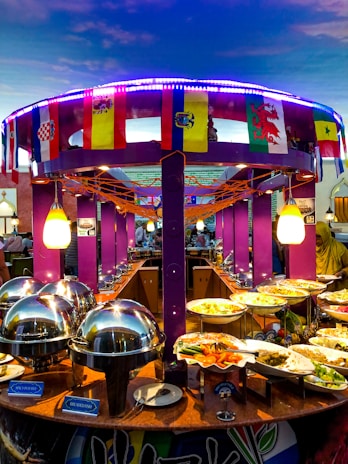 A vibrant buffet setup featuring an array of dishes including seafood and vegetables. The buffet is decorated with international flags, and the setting includes bright purple columns and string lights. Chafing dishes and labeled food containers are prominently displayed.