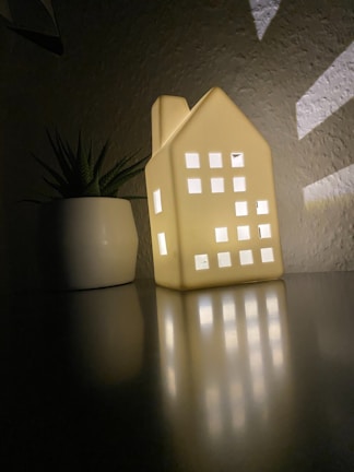 A small, house-shaped decorative light with glowing windows casting reflections on a smooth surface next to a potted plant. The setting is dimly lit, creating a cozy atmosphere.