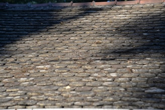 A close-up view of a stone tile roof with weathered, irregularly shaped stones. The surface features shadows and highlights, indicating the effect of sunlight and possibly overhanging trees. Small debris like twigs are scattered across the roof.