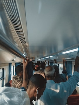 A crowded train carriage filled with standing passengers holding onto overhead rails. Most individuals are facing forward and appear to be of varying ages. Some passengers are using earphones, engaging in their own activities, while others are simply standing. The interior of the train is lit with natural light coming from windows on the left side.