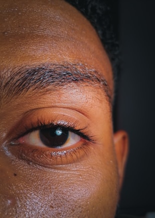 A close-up of a human eye showing detailed features such as the eyebrow, eyelid, and skin texture. The eye is open and clearly visible, displaying the iris and pupil.