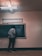 A person stands facing a chalkboard filled with mathematical equations, in a dimly lit classroom. Overhead, a fluorescent light illuminates the room, casting soft shadows. The person appears to be writing on the board, dressed casually in pants and a long-sleeved shirt.
