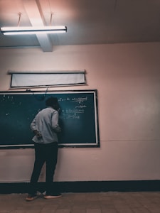 A person stands facing a chalkboard filled with mathematical equations, in a dimly lit classroom. Overhead, a fluorescent light illuminates the room, casting soft shadows. The person appears to be writing on the board, dressed casually in pants and a long-sleeved shirt.