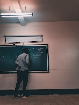 A person stands facing a chalkboard filled with mathematical equations, in a dimly lit classroom. Overhead, a fluorescent light illuminates the room, casting soft shadows. The person appears to be writing on the board, dressed casually in pants and a long-sleeved shirt.