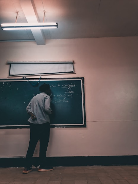 A person stands facing a chalkboard filled with mathematical equations, in a dimly lit classroom. Overhead, a fluorescent light illuminates the room, casting soft shadows. The person appears to be writing on the board, dressed casually in pants and a long-sleeved shirt.
