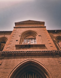 An upward view of a historic brick building with arched windows and detailed masonry. The architecture is characterized by intricate brickwork and an imposing facade that suggests an older architectural style.