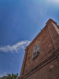 A brown brick building with a white-framed window and an air conditioning unit protruding from it, set against a bright blue sky. Wispy white clouds are scattered across the sky, and the tops of green trees are visible at the lower left corner.