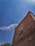 A brown brick building with a white-framed window and an air conditioning unit protruding from it, set against a bright blue sky. Wispy white clouds are scattered across the sky, and the tops of green trees are visible at the lower left corner.