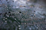 A macro shot of a circuit board highlighting intricate traces and soldered components. The focus is on the metallic elements against the green background, giving a detailed view of the tiny electronic pathways.