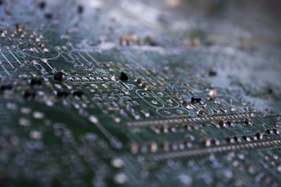 A macro shot of a circuit board highlighting intricate traces and soldered components. The focus is on the metallic elements against the green background, giving a detailed view of the tiny electronic pathways.