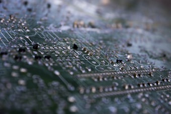 A macro shot of a circuit board highlighting intricate traces and soldered components. The focus is on the metallic elements against the green background, giving a detailed view of the tiny electronic pathways.