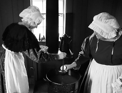 Two individuals dressed in historical attire are engaged in an indoor cooking activity. They wear bonnets and aprons typical of a 19th-century setting. One person is stirring a pot on a stove, while the other watches. The scene is illuminated by natural light coming through a window.