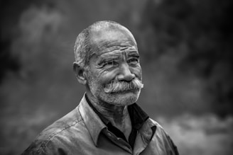 An older man with a prominent mustache and a contemplative expression is seen in a black and white photograph. His face is weathered, suggesting a life full of experiences. He wears a collared shirt, and the background is softly out of focus, adding depth to the portrait.