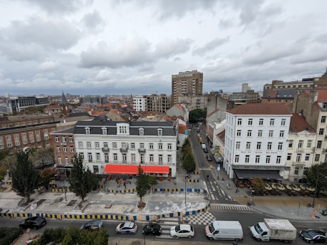 An urban scene showcasing a variety of buildings, including multi-story residential structures with both classic and modern architectural styles. The streets are lined with trees and vehicles, including cars and vans, along with several people walking and cycling. A few cafes or restaurants with outdoor seating are visible, indicating a vibrant community space. Overhead, the sky is overcast with thick gray clouds.
