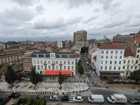 An urban scene showcasing a variety of buildings, including multi-story residential structures with both classic and modern architectural styles. The streets are lined with trees and vehicles, including cars and vans, along with several people walking and cycling. A few cafes or restaurants with outdoor seating are visible, indicating a vibrant community space. Overhead, the sky is overcast with thick gray clouds.