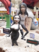 A stylized sculpture display features two cartoonish figures. The front figure is a woman with pale skin, black hair, and a black dress with a decorative neckline. The figure behind her is man with exaggerated features, long grayish hair, wearing a white tank top with printed designs and text. The scene is part of an outdoor festive setting, complete with informational signs and colorful decorations. Other elements, such as a churro stall, can be seen in the blurred background.