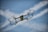 A small drone is captured in mid-flight against a backdrop of a clear blue sky with scattered white clouds. The drone is equipped with a camera and protective guards around its propellers. Its metallic body reflects some light, emphasizing its modern and sleek design.