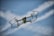A small drone is captured in mid-flight against a backdrop of a clear blue sky with scattered white clouds. The drone is equipped with a camera and protective guards around its propellers. Its metallic body reflects some light, emphasizing its modern and sleek design.