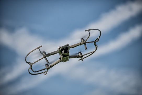A small drone is captured in mid-flight against a backdrop of a clear blue sky with scattered white clouds. The drone is equipped with a camera and protective guards around its propellers. Its metallic body reflects some light, emphasizing its modern and sleek design.